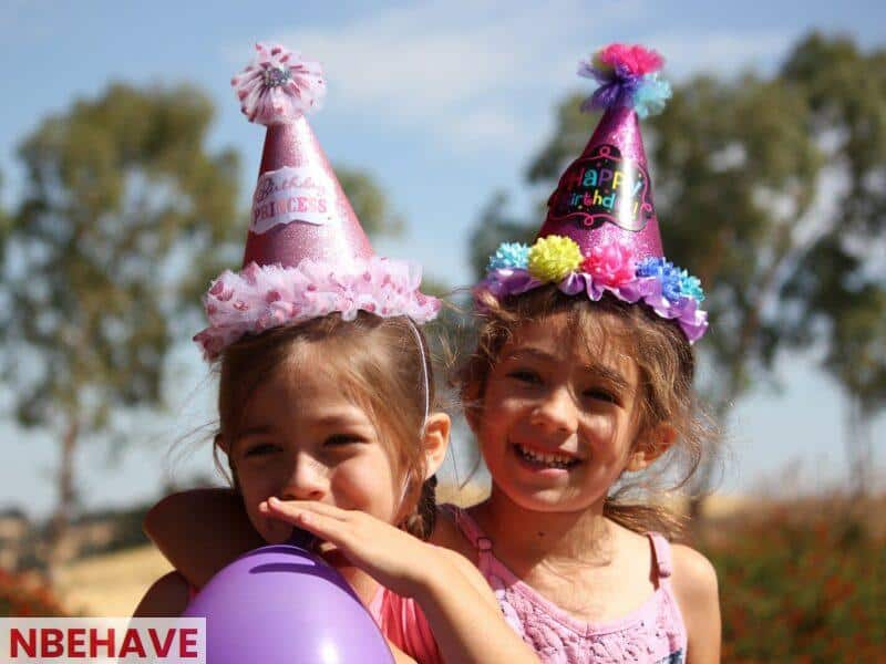 girl in left blowing purple balloon beside girl wearing pink birthday hat
