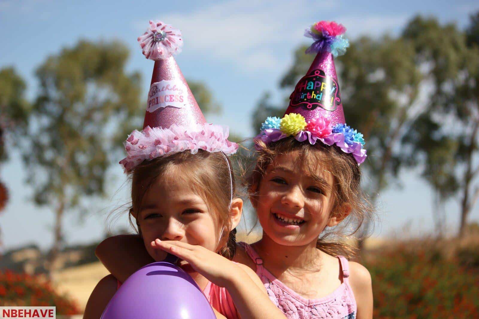 girl in left blowing purple balloon beside girl wearing pink birthday hat